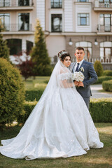 A bride and groom stand in front of a building, with the bride wearing a white dress and the groom wearing a blue suit