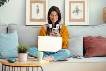 Beautiful woman working with her laptop while using mobile phone sitting on a couch at home.