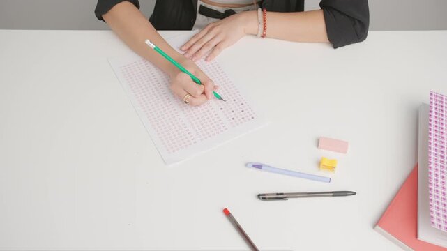 Female at desk writes correct answers in standard exam test paper with pencil. Student filling up the answers sheet with right answer bubbles, top view.
