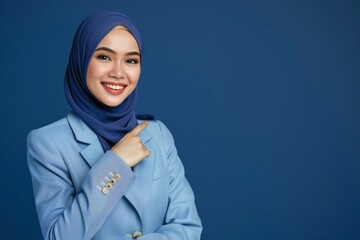  a beautiful young Malay woman with hijab smiling and pointing at something on a dark blue isolated background.