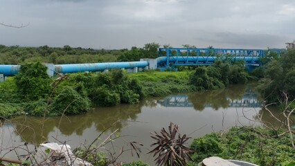 A blue-colored pipeline bridge over a river, with water beneath and a cloudy sky, emphasizing its...