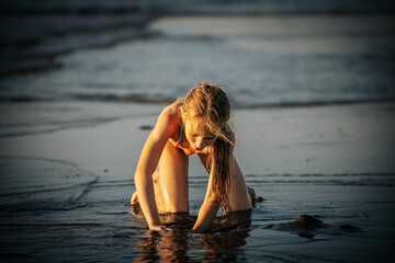 A girl kneeling and playing in the wet sand at the beach, fully engrossed in her activity. The...