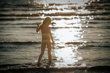Fototapeta premium A girl in a colorful swimsuit wading through sunlit waves at the beach, creating a sparkling reflection on the water. The scene captures the beauty and serenity of a beach day.