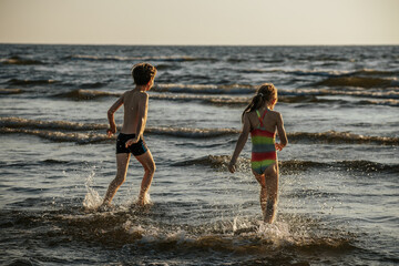 Two children are playing and splashing in the shallow water at the beach, enjoying a sunny day. The...