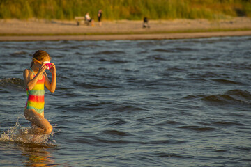 A girl in a colorful swimsuit and pink goggles playfully splashing in the waves at the beach during...