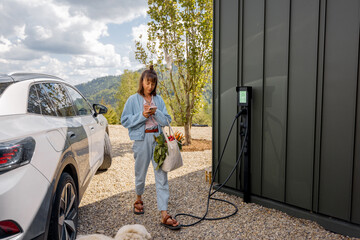 Young woman going home with groceries in bag arrived by electric car into her modern house. Concept of sustainable lifestyle, wellbeing, healthy eating and environmental care