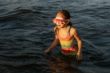 A girl in a colorful swimsuit and pink goggles playfully splashing in the waves at the beach during sunset. The image captures a joyful and energetic moment by the sea