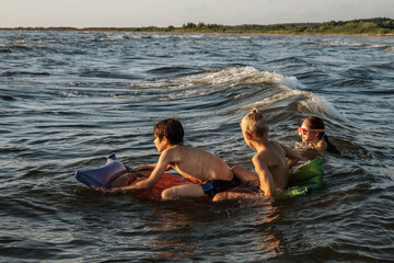 Obraz premium Three children playing in the water at the beach during sunset. The girl in a colorful swimsuit adjusts her goggles while the two boys sit on an inflatable raft. The scene captures a joyful and serene