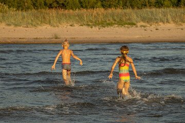 Two children are playing and splashing in the shallow water at the beach, enjoying a sunny day. The boy wears striped swim trunks, and the girl wears a colorful swimsuit and goggles.