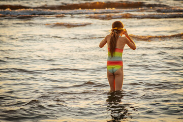 A girl in a colorful swimsuit and pink goggles playfully splashing in the waves at the beach during...