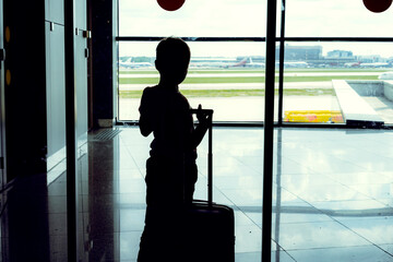 silhouette of caucasian boy with suitcase standing by the window in airport watching planes waiting...