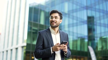 Confident businessman in suit walking near modern glass business office building holding smartphone in hands. Bearded man in glasses entrepreneur, investor, seo or manager using a mobile phone outside