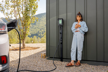 Woman stands with a smartphone and waits for electric car to be charged near a charging station at...