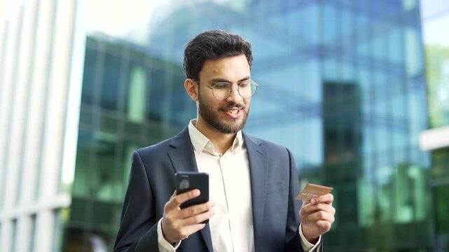 A businessman in a suit enters a credit card number on a phone while standing on the street near a business office building. Handsome bearded man shopping online, happy about good price and promotions