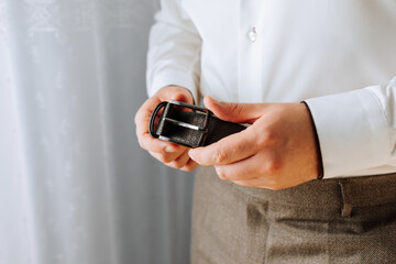 A man is wearing a belt and holding it up to his waist. The belt is brown and has a buckle. The man is dressed in a white shirt and khaki pants. Concept of formality and professionalism