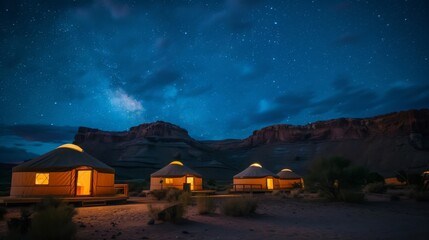 A row of lit yurts sits before a backdrop of red rock cliffs and a dark, starry sky.