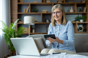 Senior woman uses calculator while working with laptop managing household budget. Smiling while paying utilities and rent payments. Concept of financial planning and budgeting.