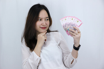 Beautiful Asian young woman holding cash money in Indonesian rupiah keeps index finger on chin and looking thoughtful, poses against white background