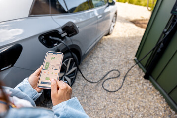 Woman using smartphone with launched application for car charging, controlling the charging process remotely. Smart technologies and app design concept