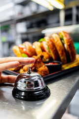 hand pressing service bell in restaurant setting, with a blurred background featuring a sandwich and side dishes, indicating prompt service. Ideal for hospitality, dining, and customer service themes.