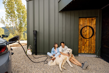 Lovely couple sitting with their dog near a modern house, electric car charges nearby. Concept of...