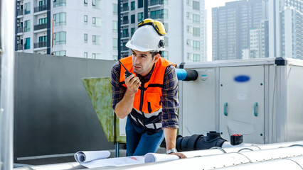 Male technical foreman inspects maintenance working in blueprint and commands with talking on radio call with colleague with to look at plumbing and electrical systems on the roof of a building