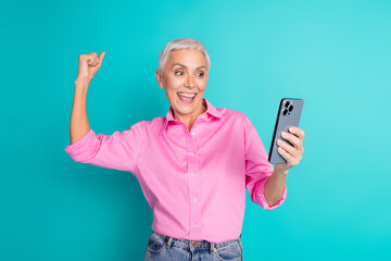 Portrait of pretty aged lady raise fist use phone wear pink shirt isolated on turquoise color background