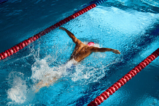 Swimmer in pink cap executes powerful butterfly stroke, his muscular form cutting through blue water, creating dynamic splash between red lane dividers. Concept of sport, preparation to competition