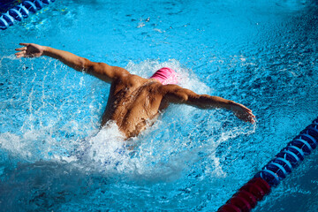 Overhead view of athlete in pink swim cap performing butterfly stroke, showcasing strength and technique as he propel through vibrant blue pool. Concept of aquatic sport, preparation to competition
