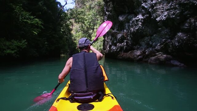 Man skillfully maneuvers kayak through calm river using paddle. Male relishes kayaking session taking in stunning views of cliffs on summer day