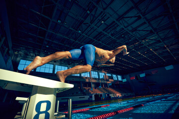 Dynamic photo of young muscular man, professional swimmer diving into pool from starting platform...