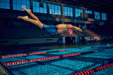 Swimmer in pink cap and blue trunks dives gracefully into indoor pool from starting platform against backdrop of empty stadium. Concept of aquatic sport, preparation to competition, energy.