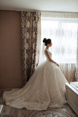 Fototapeta premium A woman in a wedding dress stands in front of a window. The dress is long and white, and the woman is wearing a veil. The room has a window with curtains, and there is a bed in the background