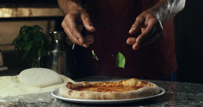 Super slow motion of pizzaman puts fresh green basil leaves falling on traditional italian dish of pizza margherita with tomato sauce and mozzarella cheese in rustic restaurant kitchen at 1000 fps.