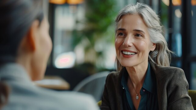 A happy senior woman listens intently to her friend, enjoying a conversation over coffee in a cozy cafe.  The image symbolizes friendship, connection, communication, and active listening.