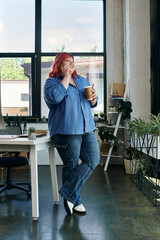 A plus size businesswoman in blue leans against a desk, enjoying coffee in a modern office.