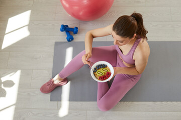 Beautiful young woman eating cereal with fruit and berries while sitting on yoga mat on floor with fit ball and dumbbells. High angle top overhead view from above. Food, sport, healthy diet concept