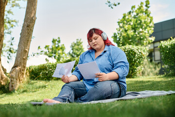 A plus size woman in a blue shirt sits on a blanket, reading papers in a lush green garden.