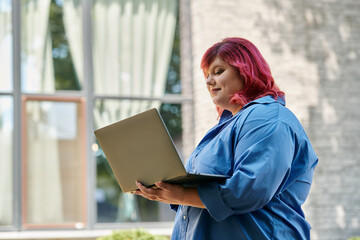 Fototapeta premium A plus size woman, wearing vibrant clothing, checks her email on a laptop outside a building.