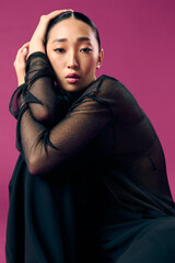 Fashion asian woman in sheer top and black pants sitting on purple background with eyes closed and...