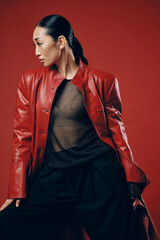Fashion asian woman in red leather jacket and black pants posing against red background with eyes...
