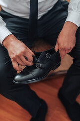 Fototapeta premium A man is sitting on a wooden floor and adjusting his shoes. The shoes are black and have a buckle on the front. The man is wearing a white shirt and black pants