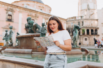 Attractive female tourist is exploring new city. Redhead happy woman holding a paper map near...