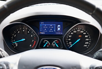 Close-up of a car dashboard with illuminated gauges and digital display showing various vehicle metrics. The image focuses on the speedometer, tachometer, fuel gauge.