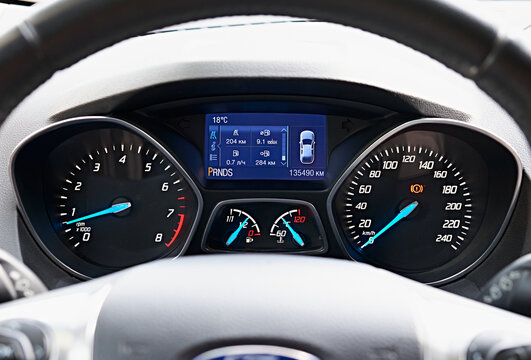Close-up of a car dashboard with illuminated gauges and digital display showing various vehicle metrics. The image focuses on the speedometer, tachometer, fuel gauge.