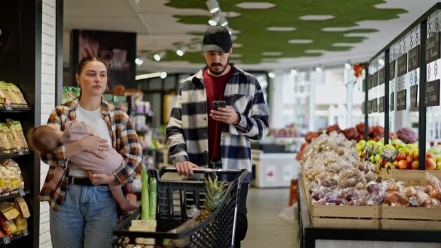 A man in a plaid shirt looks at the grocery list on his phone with his wife and small infant while shopping in a supermarket