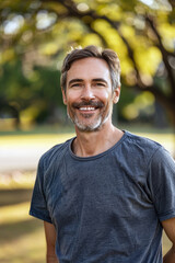 Obraz premium A mature man with gray hair and beard, wearing a gray t-shirt, standing outdoors and smiling, green blurred background