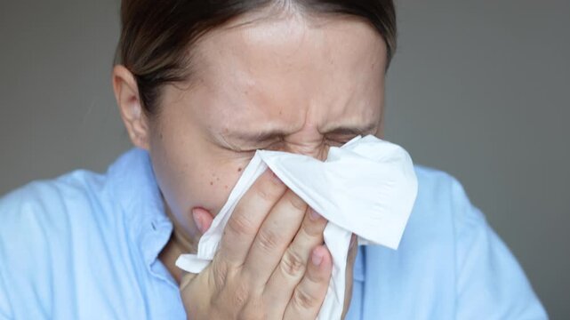 A young caucasian woman sneezing from allergy covering the nose with a facial tissue on a dark grey background. Disease, virus, flu, cold, seasonal allergy, runny nose. Slow motion