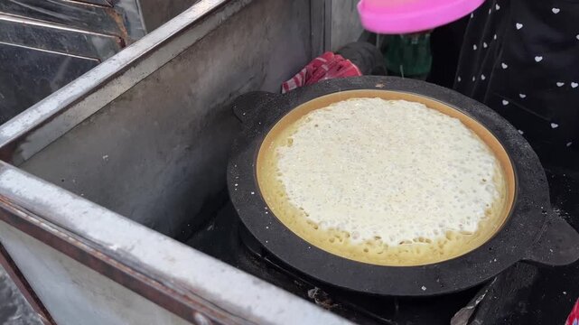 A person makes Indonesian dessert Terang Bulan, pouring batter onto a griddle and adding toppings. The process shows the dessert's golden crust and delicious fillings