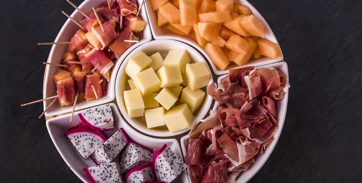 Beer snacks and tapas: cheese, jamon iberico serrano on white round plate on dark background. Food and drinks